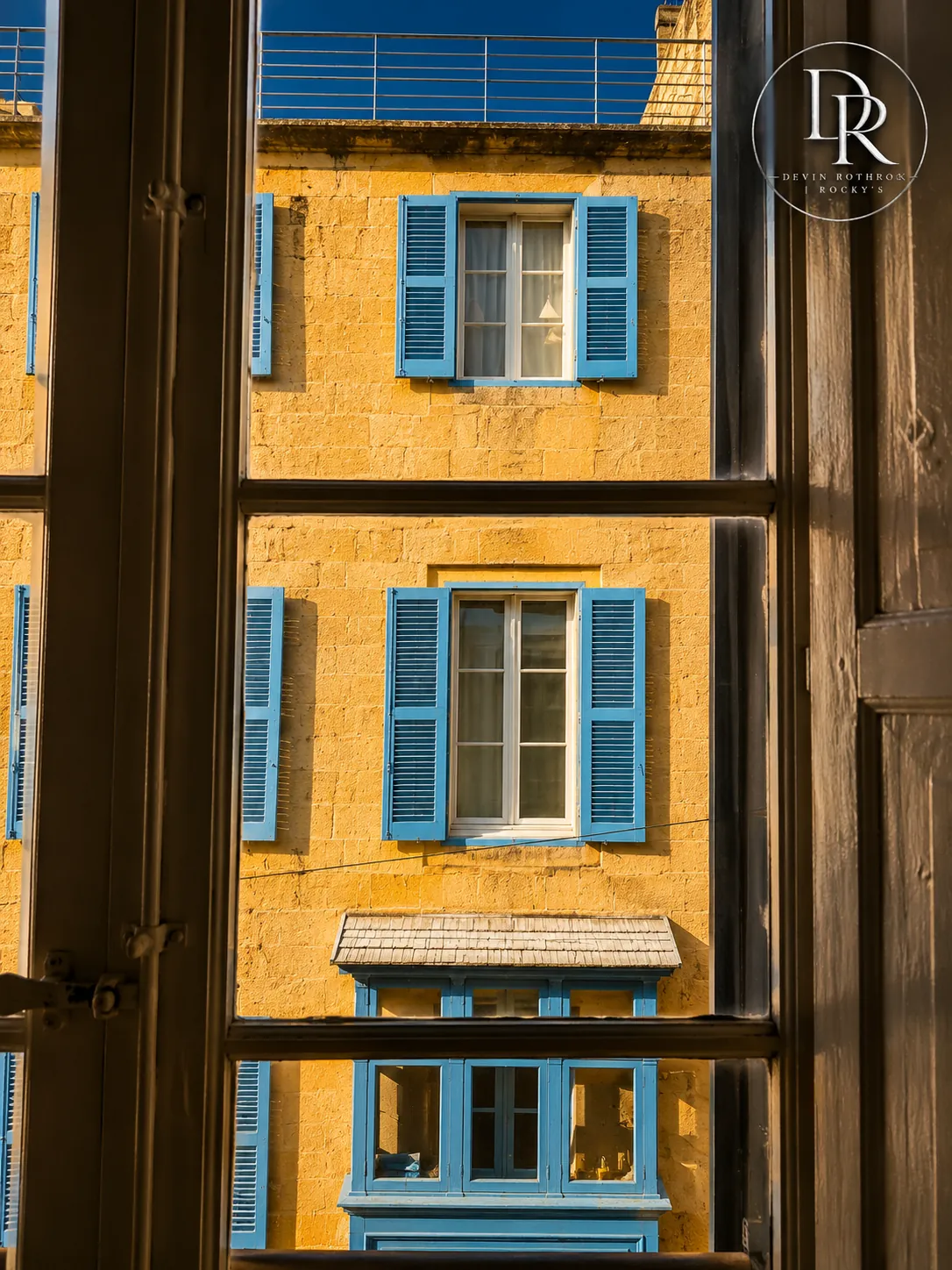 Mediterranean window view with blue shutters and golden stone, Rocky's eXp watermark